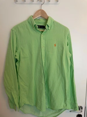 Ljusgrön skjorta från Ralph Lauren - Snygg limegrön skjorta från Ralph Lauren med klassisk button-down krage och broderad logga i orange på bröstet. Skjortan har långa ärmar och knappar framtill. Perfekt för dig som vill ha en stilren och fräsch look. Pris är ej hugget i sten. 
