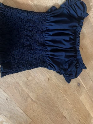Svart  smockad offshoulder topp - Trendig svart topp med smockad kropp och volangiga offshoulder-ärmar. Sitter tight över överkroppen och har en stretchig passform som framhäver figuren. Perfekt att styla med både jeans och kjol för en cool look.