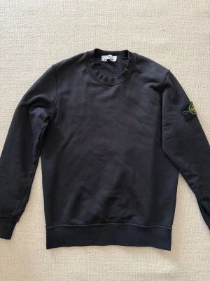 Svart sweatshirt från Stone Island - Säljer en svart sweatshirt från Stone Island med klassisk rund halsringning och ribbade muddar. Tröjan har den ikoniska Stone Island-loggan på vänster ärm. Perfekt för dig som gillar stilrena och trendiga plagg.