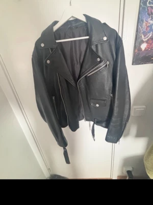 Svart klassisk skinnjacka bikerstil - Säljer en svart skinnjacka i bikerstil med breda slag, dragkedjor och axelklaffar. Jackan har fickor med dragkedja och en bälte i midjan för extra edge. Perfekt för dig som vill ha en tidlös och cool look. Från EMP shop, säljer den för den va för stor, har haft på mig på den 1 gång. 