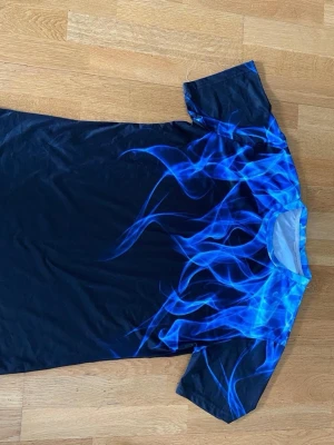 Svart t-shirt med blå eldflammor - Cool svart t-shirt med blå eldflammor som motiv över axlar och bröst. T-shirten har rund hals och korta ärmar. Materialet känns mjukt och stretchigt, troligen polyester. Perfekt för dig som vill sticka ut med en unik design.