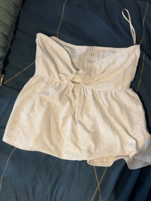 Vit axelbandslös topp från SHEIN - Säljer en söt vit axelbandslös topp från SHEIN i storlek S. Toppen har broderade hål, twistad detalj framtill och en luftig passform. Tillverkad i 95% polyester och 5% elastan, perfekt för varma dagar.