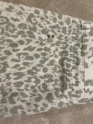 Leopardmönstrade jeans från Bershka - Säljer ett par vita jeans med grått leopardmönster från Bershka. Modellen har klassiska fem fickor, bälteshällor och knappgylf. Perfekta för dig som vill sticka ut med ett djurmönstrat plagg i garderoben.