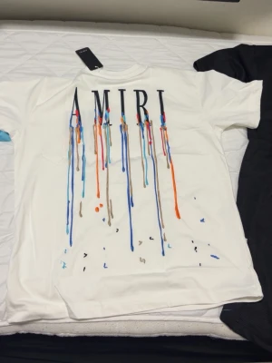 Vit AMIRI t-shirt med färgstänk - Snygg vit t-shirt från AMIRI med coolt tryck av färgstänk i blått, orange och brunt. Stor AMIRI-logga upptill på bröstet. Klassisk passform och korta ärmar, perfekt för dig som vill sticka ut med en unik design.