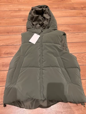 Zara Olivgrön dunväst med huva - Snygg olivgrön dunväst med dragkedja och stor huva. Västen har två snedställda fickor framtill och quiltad design. Perfekt för lager-på-lager och chill stil. Materialet är glansigt och känns mjukt.
