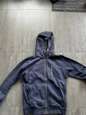 Marinblå sail racing hoodie med dragkedja - Snygg marinblå hoodie från Sail Racing med hel dragkedja framtill, huva med dragsko och en bröstficka med dragkedja. Tillverkad i mjukt bomullsmaterial och har två sidofickor. Perfekt för en avslappnad och sportig stil.