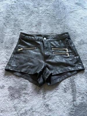 Svarta höga shorts i skinnimitation - Snygga svarta shorts i skinnimitation med hög midja och tre silverfärgade dragkedjor framtill. De har en cool och edgy look, perfekta till en statement-outfit. Materialet är syntetiskt och shortsen har en figurnära passform. Kolla alla bilder för att se alla defektet