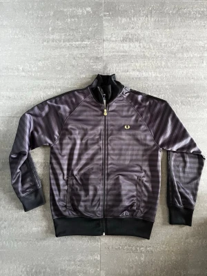 Svartgrå randig track jacket Fred Perry - Snygg svartgrå track jacket från Fred Perry med diskreta horisontella ränder och guldfärgad logga på bröstet. Dragkedja framtill, ståkrage och muddar vid ärmslut och nederkant. Två fickor fram och sportig retrostil.