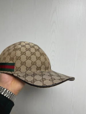 Gucci keps - (ÄKTA) Snygg beige keps från Gucci med klassiskt GG-monogram över hela kepsen och en grön-röd rand på sidan. Kepsen har böjd skärm och är tillverkad i canvasmaterial med svart insida. Perfekt för dig som vill ha en exklusiv och trendig accessoar. Ingen box, kvitto, dustbag eller något sånt medföljer Tyvär men kepsen är självklart ÄKTA!!