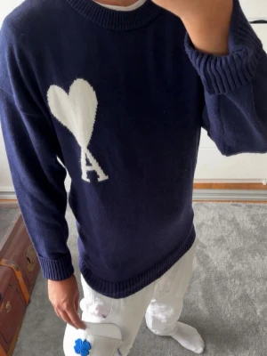 Mörkblå stickad tröja Ami Paris - Mörkblå stickad tröja från ami alexandre mattiussi med rund hals och ribbade muddar. Stor vit hjärtlogga med bokstaven A på bröstet ger en clean och trendig look. Perfekt för dig som gillar streetstyle och snygga detaljer.