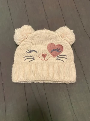 Beige stickad mössa med kattmotiv - Supersöt beige stickad mössa med två fluffiga öron på toppen. Framsidan har ett broderat kattansikte med ett rosa hjärta som ena öga och broderade morrhår. Perfekt för dig som gillar gulliga accessoarer och vill hålla dig varm.