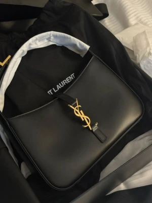 Svart axelremsväska från Saint Laurent - Stilren svart axelremsväska från Saint Laurent i slätt läder med ikoniskt YSL-monogram i guld framtill. Väskan har en justerbar rem och minimalistisk design som ger en lyxig känsla. Perfekt för dig som gillar klassiska och trendiga accessoarer.