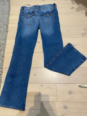 Blå bootcut jeans med fickdetaljer - Säljer ett par klassiska blå bootcut jeans med snygga fickdetaljer bak och ljusa slitningar framtill. Jeansen har normal midja och raka ben med lätt utsvängning. Perfekta till en avslappnad och trendig look. Storlek 170 men skulle säga att dom passar bättre för dom som är runt 160