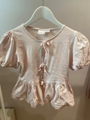Ljusrosa blus med puffärm och knyt - Supersöt ljusrosa blus med puffiga korta ärmar och tre knyt framtill. Blusen har volang nedtill och är gjord i mjuk bomull, perfekt för en trendig och avslappnad look.
