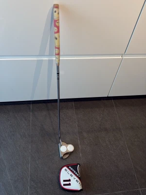 Odyssey Eleven White  Hot VÄNSTER putter - Vänster Golfputter från Odyssey, modell Eleven White Hot. Klubban har ett silverfärgat klubbhuvud med två vita cirklar och svart text. Greppet är mönstrat i beige och rött med smileys. Headcover medföljer och har svart, vitt och rött tryck.