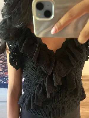 Zara topp - Snygg svart virkad topp med volangdetaljer framtill och längs ärmarna. Toppen är ärmlös och har en v-ringad halsringning. Perfekt för dig som vill ha en unik och trendig look med extra detaljer.