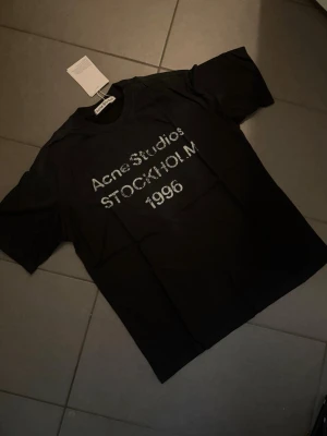 Svart Acne Studios t-shirt M - Säljer en svart t-shirt från Acne Studios med tryck i vitt framtill där det står 'Acne Studios STOCKHOLM 1996'. Klassisk rund hals och korta ärmar. Perfekt för dig som gillar stilrena och trendiga plagg.