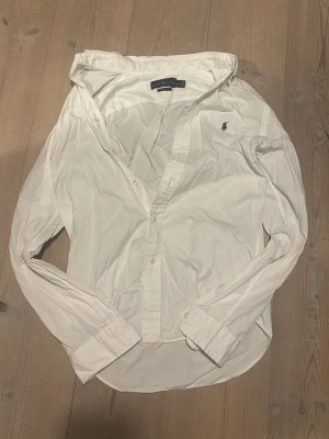 Vit skjorta från Ralph Lauren - Klassisk vit skjorta från Ralph Lauren med lång ärm och knappar framtill. Skjortan har den ikoniska broderade loggan i marinblått på bröstet och är tillverkad i bomull. Perfekt till både jeans och chinos.