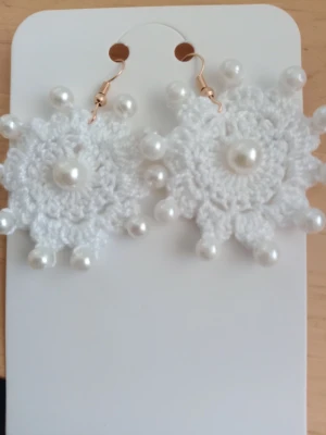 Vita virkade örhängen med pärlor - Unika handgjorda örhängen i vit virkad design med dekorativa vita pärlor runt kanten och i mitten. Örhängena har krokar i guldfärg och en snöfligeliknande form som verkligen sticker ut. Perfekta för dig som vill ha något annorlunda och kreativt.