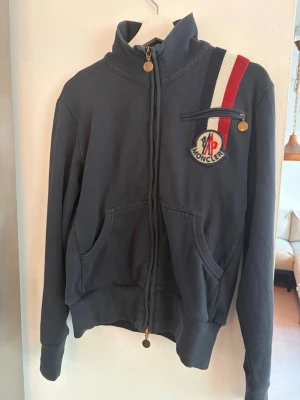 Mörkblå Moncler hoodie med dragkedja - Säljer en mörkblå hoodie från Moncler med hel dragkedja, finns det litet hål över fram fickorna som knappt syns och ett hål vid armbågen. Storlek M passar S