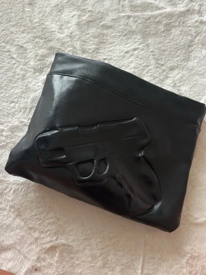 Svart kuvertväska med pistolmotiv - Unik svart kuvertväska  med ett upphöjt pistolmotiv på framsidan. Väskan har en stilren och edgy design som verkligen sticker ut. Perfekt för dig som vill ha en accessoar med attityd och cool detalj.