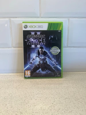 Star Wars The Force Unleashed Xbox 360 - Star Wars: The Force Unleashed till Xbox 360. Actionfyllt Star Wars-spel där du spelar som en kraftfull karaktär med episka krafter. Omslaget är svart och blått med ikoniska Star Wars-motiv. Spelfodral i plast med klassisk grön Xbox 360-design. Fungerar toppen!