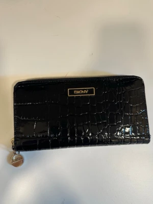 Svart plånbok i krokopräglat skinn DKNY - Snygg svart plånbok från DKNY i glansigt krokopräglat skinn. Plånboken har guldfärgade detaljer, dragkedja runtom och en metallogo framtill. Perfekt för dig som vill ha en stilren och trendig accessoar.