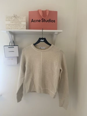 Beige stickad tröja från ZARA - Snygg beige stickad tröja från ZARA med rund halsringning och en unik vit söm som går längs framsidan. Tröjan har långa ärmar och ribbade muddar vid ärmslut och nederkant. Perfekt för lager på lager och enkel att matcha med olika stilar.