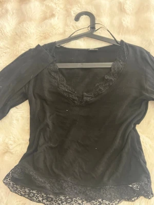 Svart spetsdetaljerad topp Gina Tricot - Svart topp från Gina Tricot med v-ringning och spetsdetaljer vid halsringning och nederkant. Tillverkad i ett mjukt material med trekvartsärm och figurnära passform. Perfekt för dig som gillar en feminin och stilren look.
