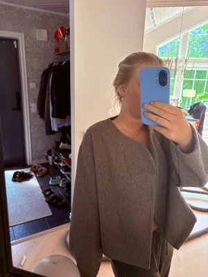 Grå cropped cardiganjacka - Snygg grå cardiganjacka med rak, boxig passform och rundad halsringning. Jackan är croppad och har långa ärmar utan synliga knappar eller dragkedja. Materialet ser ut att vara mjukt och lite tjockare, perfekt för lager på lager.
