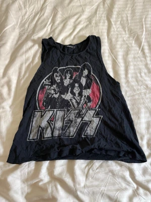 Svart KISS linnetopp från Gina Tricot - Cool svart linnetopp från Gina Tricot med stort KISS-tryck framtill. Toppen är ärmlös och har en avslappnad passform, perfekt för dig som gillar bandtröjor och rockstil. Materialet är mjukt och bekvämt bomull.