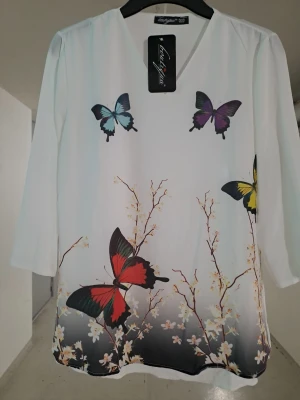 Vit blus med fjärilar och blommor - Vit blus från Lady Spirit med v-ringning och trekvartsärm. Framsidan pryds av färgglada fjärilar och blommor i rött, blått, gult och lila. Materialet känns lätt och luftigt, perfekt för en fräsch look. Passar dig som gillar färgstarka detaljer och naturinspirerade mönster.