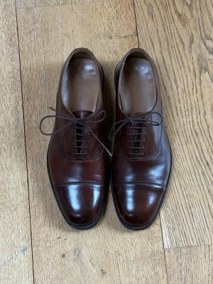 Lindeberg Hopper Cap Toe Oxford | Cognac | Made in Italy | Stl UK 7 / EU 40–41 - Klassisk cap-toe oxford från J.Lindeberg i cognacfärgat kalvskinn. Tillverkad i Italien med lädersula – ovanligt i det här prissegmentet och gör dem sulningsbara för många års fortsatt användning.  Skick: Begagnat men välvårdat. Ovandelen och formen i mycket gott skick. Normalt slitage på lädersulan. Liten repa på tåhättan som endast syns på nära håll (se bild).  Passar till kostym, chinos eller mörkare jeans.  Nypris 2 500 kr