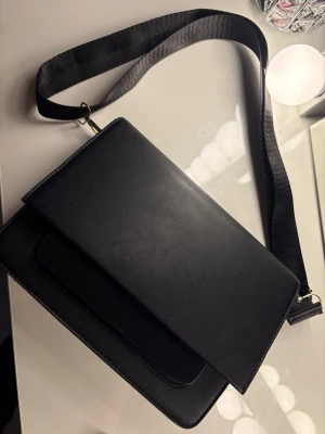 Svart axelväska i stilren design - Snygg svart axelväska från Kappahl med slät yta och bred axelrem. Väskan har ett lock med magnetknäppning och en praktisk innerficka. 