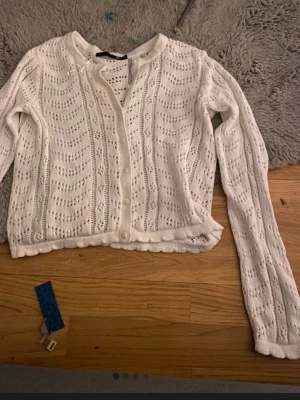 Vit virkad kofta med knappar - Söt vit stickad kofta med virkade detaljer och knappar framtill. Kortare modell med rund halsringning och långa ärmar. Perfekt att slänga över en topp för en chill look.