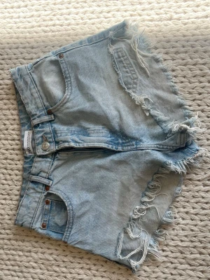 Ljusblå denimshorts med fransar - Säljer ett par ljusblå denimshorts med råa, fransiga kanter och klassisk femficksdesign. Shortsen har hög midja och en avslappnad passform, perfekta för varma dagar. 