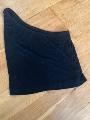 Svart oneshoulder topp från SHEIN - Snygg svart oneshoulder topp från SHEIN i ribbat material. Tillverkad av 60% viskos, 35% polyester och 5% elastan för en stretchig och bekväm passform. Perfekt till sommarens outfits och enkel att styla.