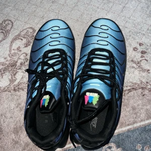 Blå Nike Air Max Plus sneakers - Säljer ett par blå Nike Air Max Plus sneakers med svarta detaljer och vågigt mönster på ovandelen. Skorna har synlig Air-sula, svart yttersula och färgglad logga på plösen. Materialet är mesh och syntet, och modellen har en sportig vibe.