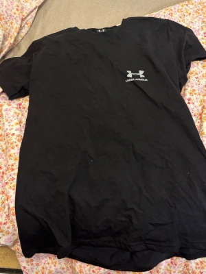 Svart t-shirt från Under Armour - Svart t-shirt från Under Armour med klassisk passform och diskret logga på bröstet. Tillverkad i mjuk bomull som känns skön mot huden. Perfekt för en sportig och avslappnad stil.