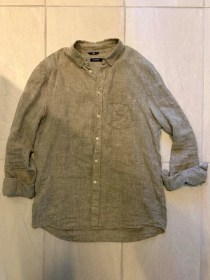 Linneskjorta J.Lindeberg herr  - Snygg beige skjorta i linne med klassisk krage och knäppning framtill. Skjortan har en bröstficka och långa ärmar som kan vikas upp för en mer avslappnad look. Perfekt för dig som gillar en enkel och stilren stil.
