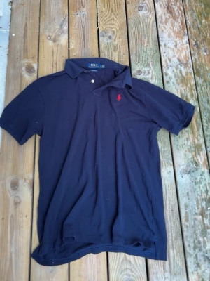 Mörkblå pikétröja från Polo Ralph Lauren - Klassisk mörkblå pikétröja från Polo Ralph Lauren med korta ärmar och krage. Tröjan har två knappar vid halsen och den ikoniska röda logotypen broderad på bröstet. Tillverkad i mjukt bomullsmaterial som är skönt mot huden.