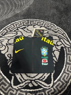 Svart Brasil Nike träningsjacka - Snygg svart träningsjacka från Nike med Brasiliens landslagslogga på bröstet. Jackan har gula och orangea tryck med sponsorloggor, hel dragkedja och två sidofickor. Tillverkad i lätt och funktionellt material, perfekt för träning eller sport.