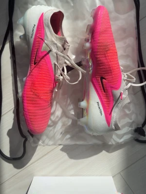 Nike Phantom rosa fotbollsskor - Säljer ett par Nike Phantom fotbollsskor i en snygg rosa och vit färgkombination med svarta detaljer. Skorna har en stickad ovandel och snörning för bra passform samt en sula med dobbar i transparent och regnbågsskimrande finish. Perfekta för dig som vill sticka ut på planen!