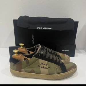 Saint Laurent camo sneakers - Snygga Saint Laurent sneakers i camomönstrad canvas med svarta detaljer och snörning. Skorna har en låg silhuett och Saint Laurent-loggan broderad på sidan. Perfekta för dig som vill sticka ut.