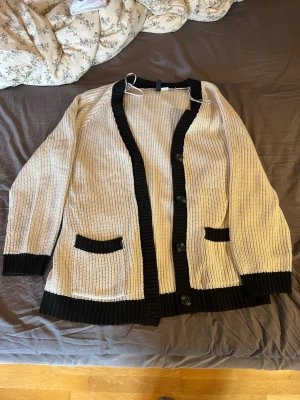 Beige kofta med svarta detaljer Divided - Säljer en beige stickad kofta från Divided med svarta ribbade kanter, knappar framtill och två fickor. Koftan har lång ärm och är perfekt för lager på lager. Materialet är mjukt och skönt, passar till många olika stilar.Är i storlek M men passar även L