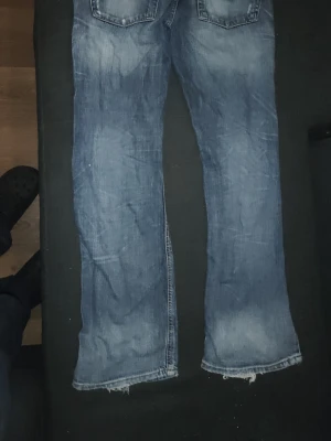 Blå slitna bootcut jeans - Ett par blå bootcut jeans med slitningar och råa kanter nertill. Jeansen har klassiska fem fickor och tydliga slitningar på både fram- och baksida för en avslappnad look. Tillverkade i denim med en tvättad finish.