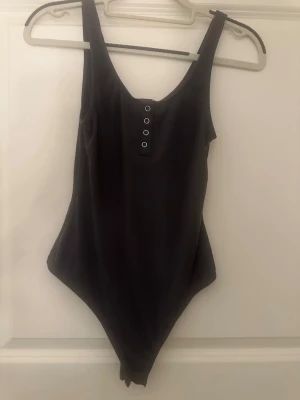Svart ribbad body med knappar - Snygg svart body i ribbat tyg med rundad urringning och knappdetaljer framtill. Modellen har breda axelband och hög skärning vid benen. Perfekt att styla till jeans eller kjol för en trendig look.