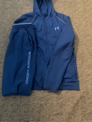 Blått träningsset från Under Armour - Säljer ett blått träningsset från Under Armour med jacka och byxor. Jackan har huva, dragkedja och logga på bröstet. Byxorna har Under Armour-tryck längs benet. Perfekt för löpning eller gym. Materialet är lätt och funktionellt.