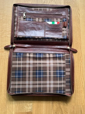 Brun portfölj i läder med rutigt foder - Snygg portfölj i brunt italienskt läder med rutigt tygfoder i beige, blått och brunt. Portföljen har flera fack för kort, dokument och dragkedja för småsaker. Stängs med dragkedja runt om och har klassisk, stilren design. Köpte den för 185 euro denna sommaren men kommer inte komma till användning