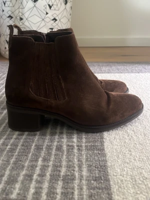 Bruna Chelsea boots i mocka - Snygga Chelsea boots i mörkbrun mocka med klassisk resår på sidorna och låg klack. Bootsens form är stilren med rundad tå och dragflik bak för enkel påtagning. Perfekta för dig som vill ha en tidlös och trendig look.